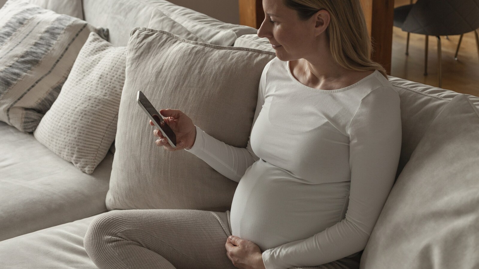 Gravid kvinna i beige kläder sitter i soffa och tittar i mobilen