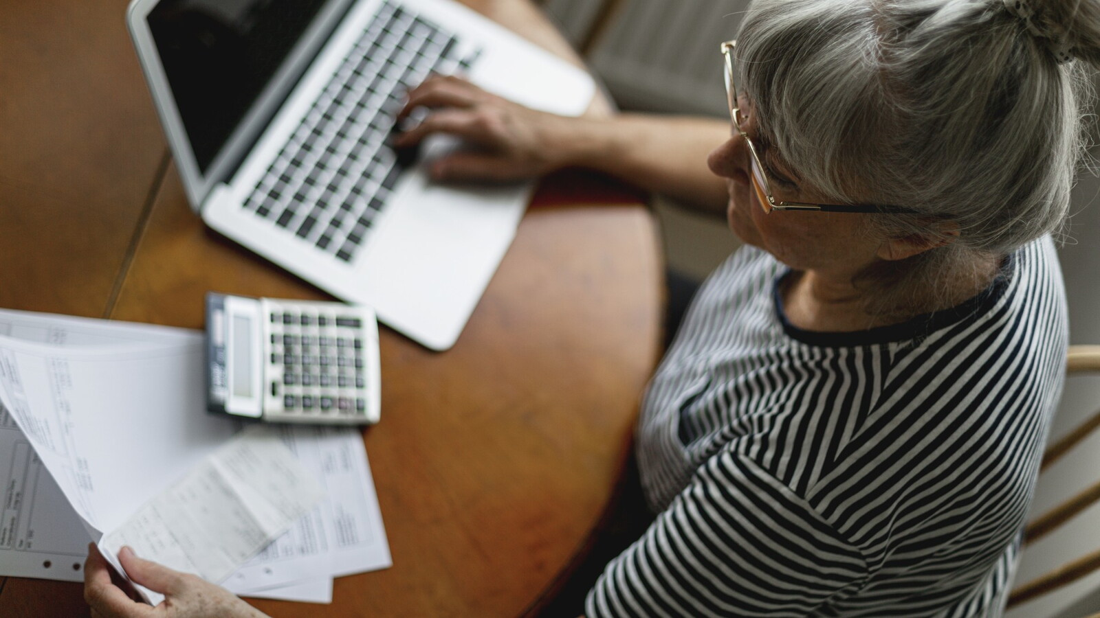 Äldre kvinna sitter vid bord med laptop och miniräknare